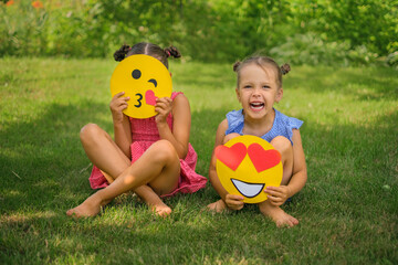 Hilariously laughing children are sitting in the park on the lawn with various paper emoticon faces. Smile face with hearts and emoji sending an air kiss