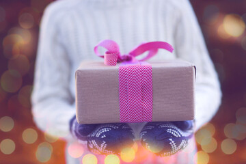 Wall Mural - close-up of nicely wrapped christmas gift being held by the kid in mittens by christmas tree with lights, toned image