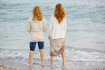 Red-haired elderly mother and her grown-up daughter are walking along the seashore together. Happy meeting of a mother and her grown-up child. Family relations between adult children and parents