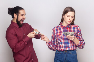 Portrait of serious rich confident woman and man gigolo standing together, wife giving money to her husband with indifferent face. Indoor studio shot isolated on gray background.