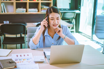 Wall Mural - Business asian woman sitting at her desk with laptop using mobile phone chatting with customers or giving financial advice.