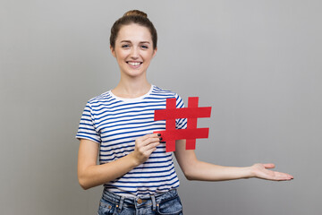 Wall Mural - Portrait of woman in striped T-shirt holding hashtag and presenting at copy space on her palm, empty place for idea presentation, product advertising. Indoor studio shot isolated on gray background.