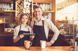© Designpics - Professional barista. Young woman and man in aprons looking at camera, smiling and standing at bar counter. Woman using tablet computer