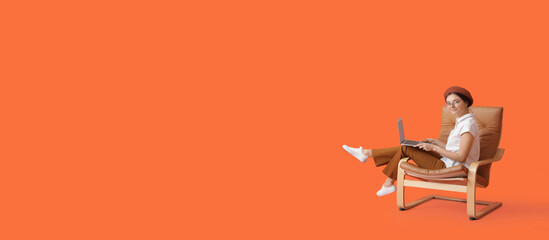 Beautiful young woman with laptop sitting in armchair on orange background with space for text