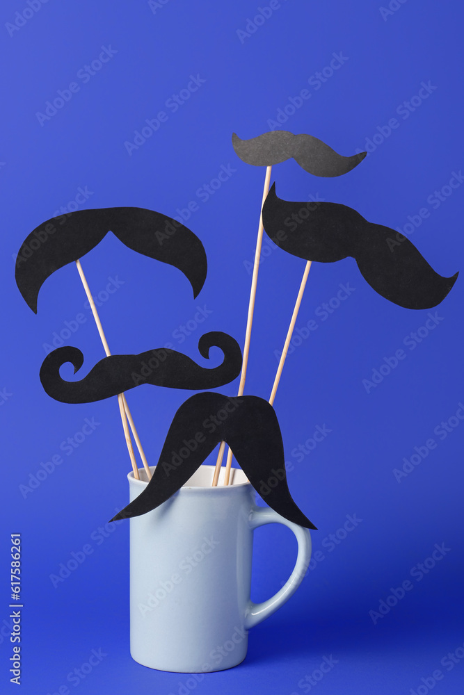 Wooden sticks with different paper mustaches in cup against blue background
