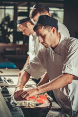 Wall Mural - Three young chefs dressed in white uniform decorate ready dish in restaurant. They are working on maki rolls. Preparing traditional japanese sushi set in interior of modern professional kitchen