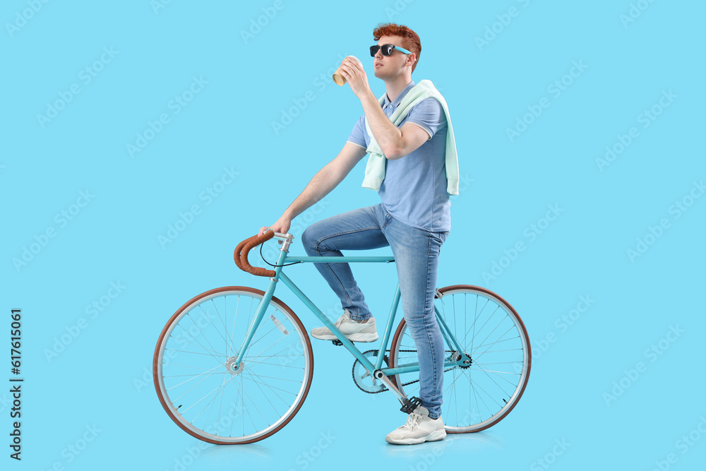 Young redhead man with bicycle and cup of coffee on blue background