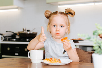 Sad little caucasian girl eating fast food while sitting at the table in the kitchen and showing like gesture