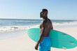 © wavebreak3 - Fit african american man holding surfboard on sunny beach, looking away