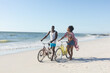 © wavebreak3 - Happy african american couple walking with bikes on sunny beach by sea