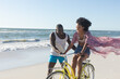 © wavebreak3 - Happy african american couple having fun walking and riding bike on sunny beach by sea, copy space