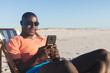© wavebreak3 - Happy african american man in sunglasses sitting in deckchair using smartphone on sunny beach