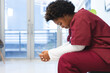 Tired african american female doctor wearing scrubs, sitting on chair in corridor at hospital