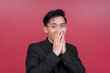 © Mdv Edwards - Portrait of a shocked young man wearing glasses with both hands on his mouth. Isolated on a red background.