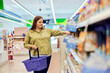 A plus-size woman holding a  basket and choosing groceries from a shelf.
