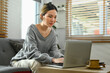 © Prathankarnpap - Asian woman in casual clothes sitting on couch and using laptop. People, technology and lifestyle concept