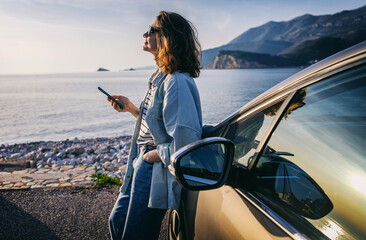 young woman traveler istanding by her car during summer sea holiday on the beach at sunset