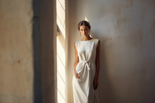 A Beautiful Model In A White Dress Stands Near A Sandstone Wall In The Sun.
