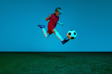 Wall Mural - Studio shot of little boy, soccer player in sports uniform jumping with football ball over dark blue background in neon. Concept of sport, health, action, ad