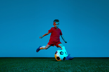 Wall Mural - One sportive child, boy in red uniform playing, training football over dark blue background in neon. Concept of action, sportive lifestyle, team game, health, energy