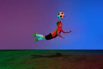 Wall Mural - Active, energetic school age boy, football player training with soccer ball over dark blue background in neon. Concept of team sport, health, action, ad