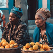 © ADDICTIVE STOCK - Generative AI illustration of sad senior African female vendors with headwraps looking away while sitting in local market with fruits