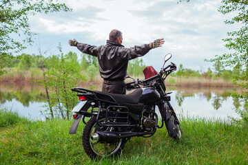 Back view of happy senior biker raising arms enjoying river landscape after riding motobike. Traveling by motorcycle