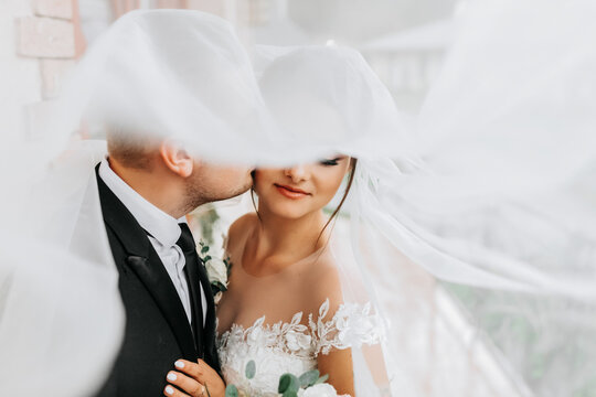 Stylish newlywed European couple. Smiling bride in a white dress. The groom, dressed in a classic black suit, white shirt, kisses the bride on the cheek under the veil. Wedding in nature