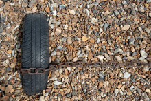 Rusty Chain Fastened On An Old Black Vehicle Wheel. Beach Gravel Background.  Boat Moored In The Shore