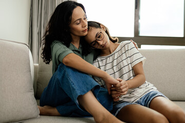 Caring mother comforting her teenage daughter, both sitting on the couch at home, sharing a tender and supportive moment