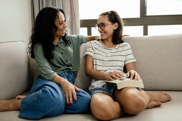 A mother and her teenage daughter laughing happily on the sofa after a satisfying read, the girl holding the book on her leg.