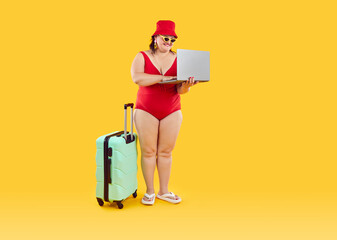 Wall Mural - Happy funny fat woman in red hat and swimsuit with suitcase getting ready for holiday trip booking tickets online via laptop isolated on studio yellow background. Vacation and summer journey concept.