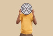 Unknown ethnic man covers his face with darts board, which symbolizes achievement of goals. Focused man in casual clothes holding darts board isolated on light beige background. Targeting concept.