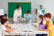 © Studio Romantic - Young smiling teacher is engaged with student standing at table against the background of blackboard, other children are laughing while sitting at the table and making notes in their school notebooks.