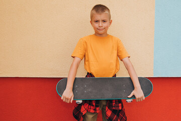 Outdoor activities for children. A cheerful young boy in bright clothes holding a skateboard against the backdrop of a colorful wall. Caucasian schoolboy on a walk. Front view