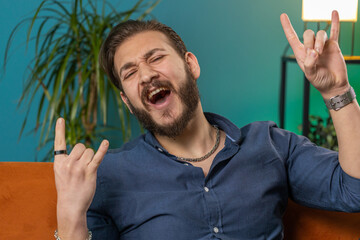 Overjoyed delighted young lebanese man showing rock n roll gesture by hands, cool sign, shouting yeah with crazy expression, dancing. Arabian guy emotionally rejoicing in success at home room couch