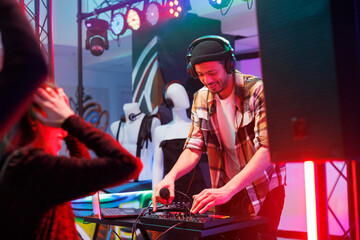 Dj in headphones using jog wheel and knobs while mixing sound with controller on nightclub stage. Young man musician playing electronic music during disco party in dark club