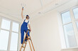 Worker in uniform painting ceiling with roller on stepladder indoors, low angle view