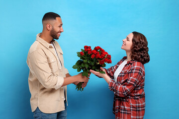 Wall Mural - Boyfriend presenting bouquet of red roses to his girlfriend on light blue background. Valentine's day celebration