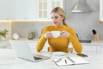 Poster - Home workplace. Woman with cup of hot drink looking at laptop at marble desk in kitchen