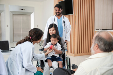 Happy young female pediatric doctor in uniform teasing little boy in wheelchair for medical exam at outpatient clinic hospital, people public health care checkup, and appointment visit.