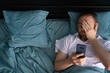 © Andrii Lysenko - Confused man looking on mobile phone lying in bed at home in the morning. Oversleeping concept.