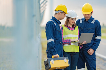 Wall Mural - A team of 2 male engineers and an Asian female supervisor working on laptops standing near the fence of an industrial factory. Put on the mechanic's uniform, vest, helmet and toolbox.