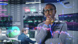 © Framestock - African American investment specialist analyzes real-time stocks in office. 3D graphics of cryptocurrency charts on glass wall. Futuristic VFX animation. Employees work on computers on background.