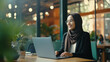 Middle Eastern Muslim woman wearing a hijab working on a laptop in a modern office or cafe on a video call