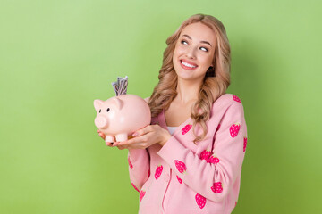 Poster - Photo of young lady blonde curly business lady hold piggy bank collect banknotes dollars look dreamy isolated on green color background