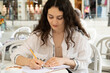 © Zarina Lukash - Beautiful young woman in a cafe sitting and writing with a pen in a notebook. Plans for the day, wish list, life diary