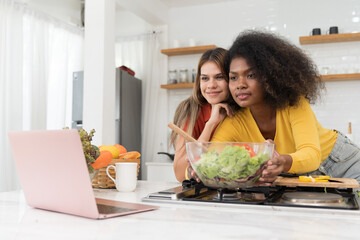 Wall Mural - Happy two young women looking laptop computer during cooking together in kitchen room at home. Two young diverse lesbian women spending time together. Diversity, LGBT and gender identity concept