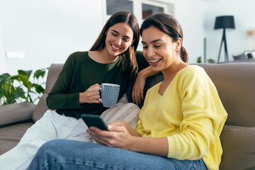 Wall Mural - Two smiling young women talking while watching smartphone sitting on couch in the living room at home.