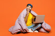 Full body young IT man wear pyjamas jam sleep eye mask sit wrapped in duvet rest relax at home work hold use laptop pc computer look aside isolated on plain orange background studio Night nap concept